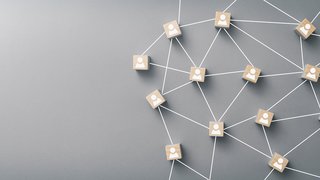 Mehrere Holzklötzchen mit stilisierten Personensymbolen sind auf einem grauen Hintergrund durch weiße Linien miteinander verbunden. Die Anordnung symbolisiert ein Netzwerk oder ein soziales bzw. berufliches Beziehungsgeflecht. Links im Bild ist viel leerer Raum.